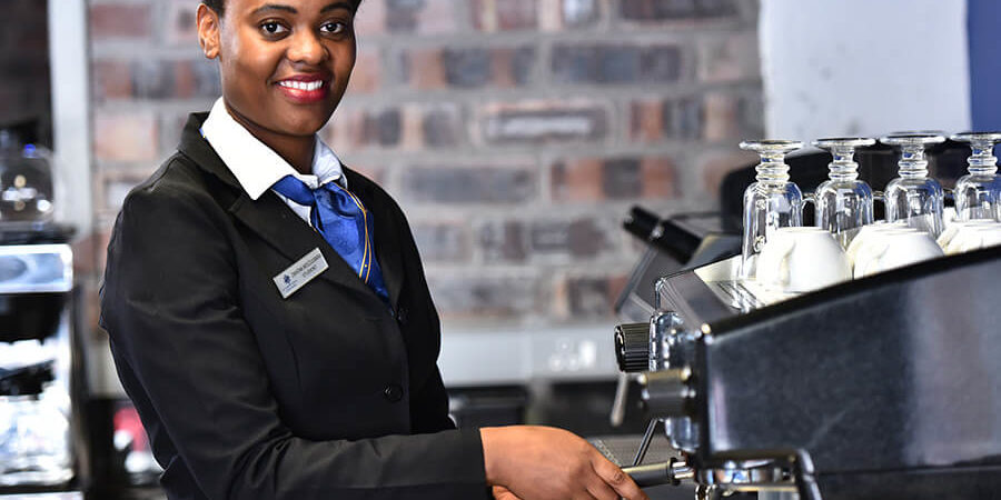 A barista in a black uniform operating an espresso machine in a café setting. - International Hotel School