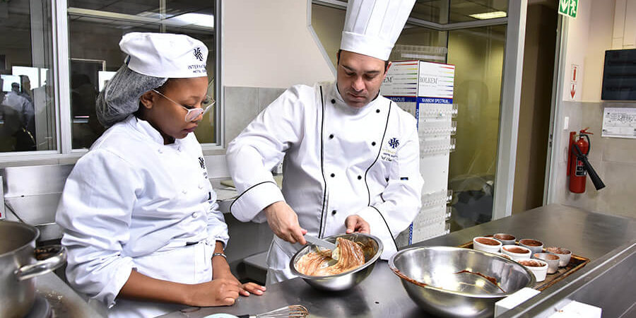 Chef demonstrating cooking techniques to a student in a professional kitchen setting. - International Hotel School