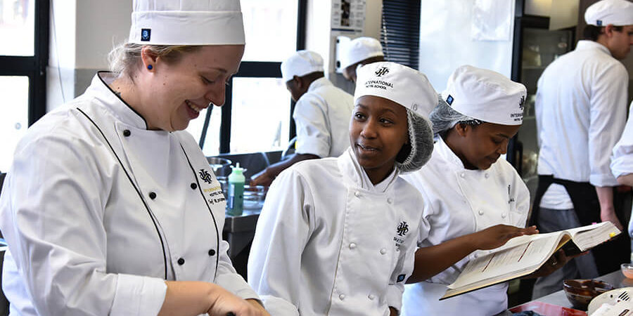 Culinary students in a professional kitchen, engaged in cooking activities with chef uniforms and hats. - International Hotel School