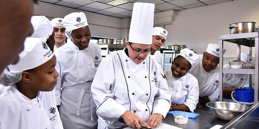 Chef instructor demonstrating knife skills to culinary students in a kitchen setting. - International Hotel School
