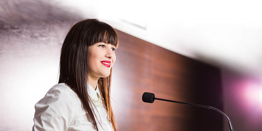 A woman with long dark hair speaking at a podium during a conference event. - International Hotel School
