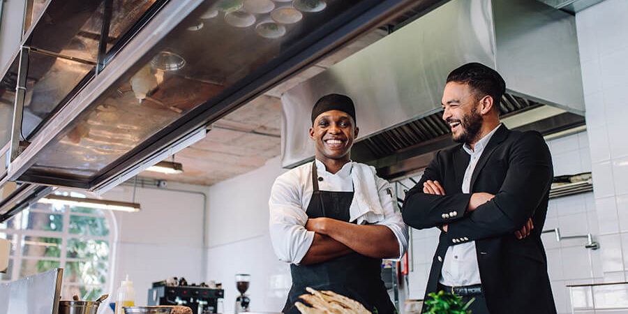 Two men smiling in a kitchen, one wearing a chef's uniform and the other in a suit. - International Hotel School