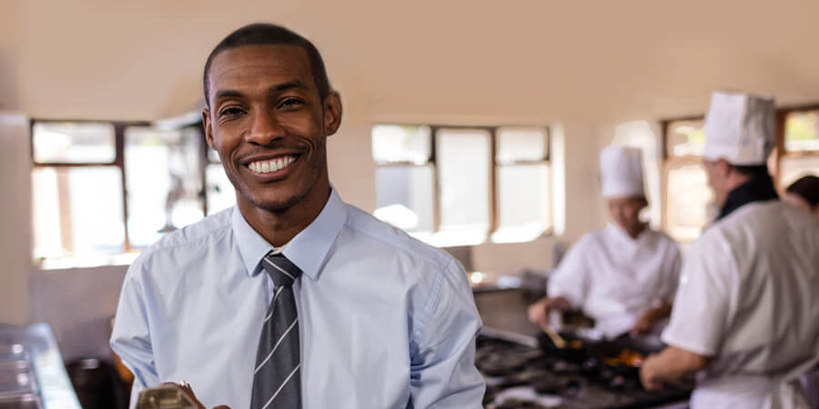 A man in a blue shirt and tie holding a clipboard in a kitchen setting with chefs in the background. - International Hotel School
