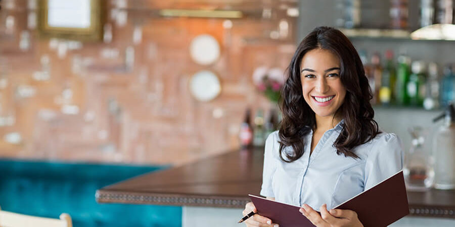 A female hospitality professional smiling while holding a menu in a restaurant setting. - International Hotel School