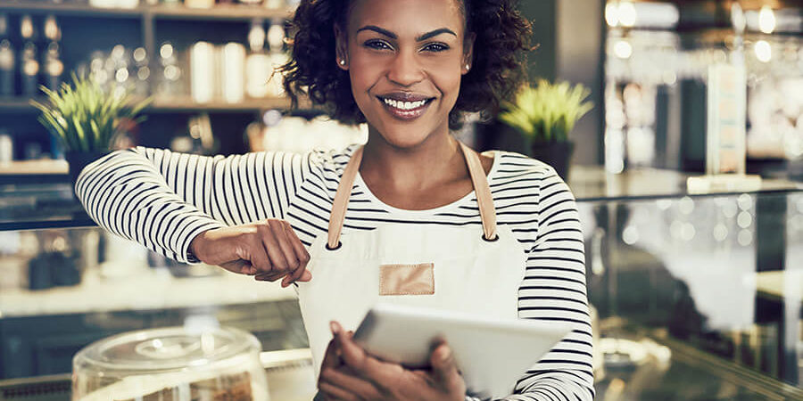 A woman wearing a striped shirt and apron, smiling while holding a tablet in a café setting. - International Hotel School
