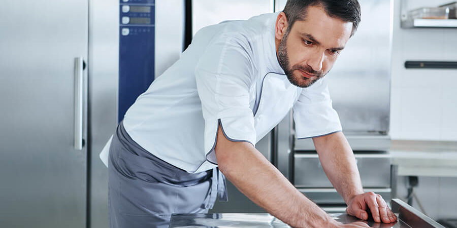 A chef in a white uniform cleaning a stainless steel surface in a kitchen. - International Hotel School