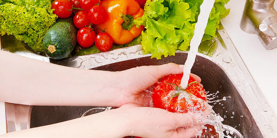 Hands washing a ripe tomato under running water in a kitchen sink with various vegetables nearby. - International Hotel School