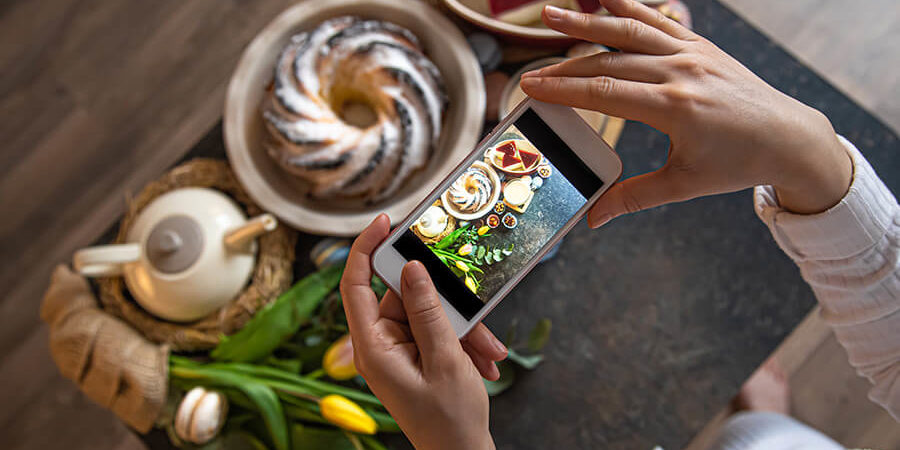 Individual using a smartphone to photograph a dessert and floral arrangement on a table. - International Hotel School