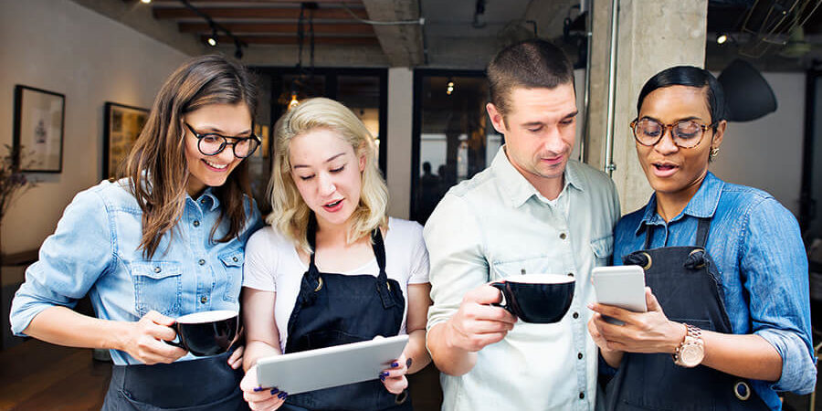 Four individuals in aprons discussing while holding coffee cups and a tablet in a casual setting. - International Hotel School