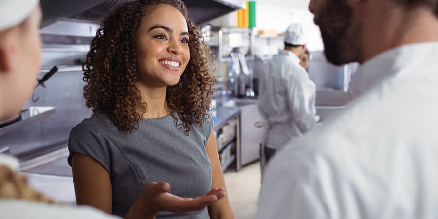 A woman with curly hair engaging in conversation with two chefs in a professional kitchen setting. - International Hotel School