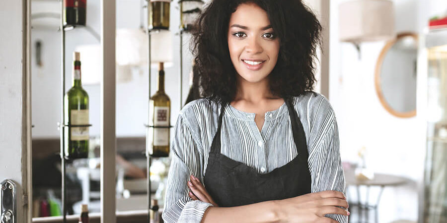 A woman wearing an apron stands with crossed arms, smiling in a hospitality setting. - International Hotel School