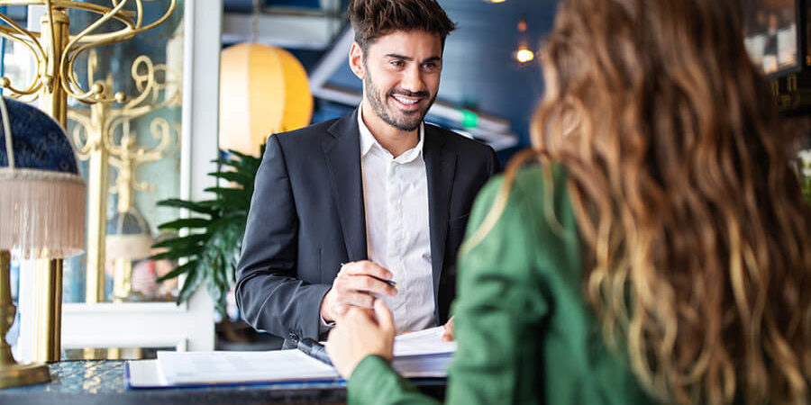 A hotel receptionist in a suit interacting with a guest at the front desk. - International Hotel School