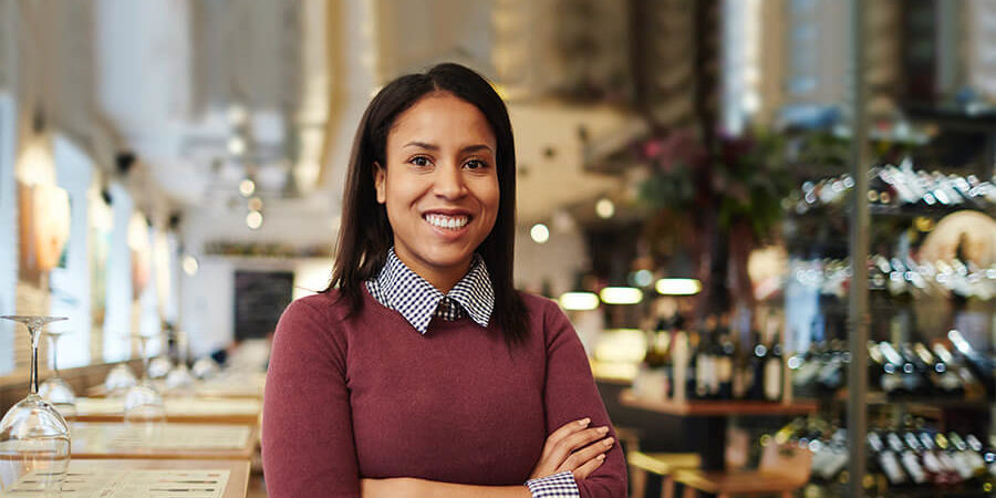 A woman with crossed arms smiling in a restaurant environment with wine bottles in the background. - International Hotel School