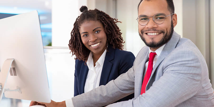 Two professionals, a man in a gray suit and a woman in a navy blazer, smiling at a computer in a modern office. - International Hotel School