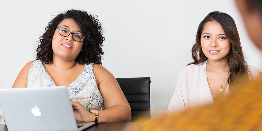 Two women seated at a table with a laptop during a meeting, one looking at the camera. - International Hotel School