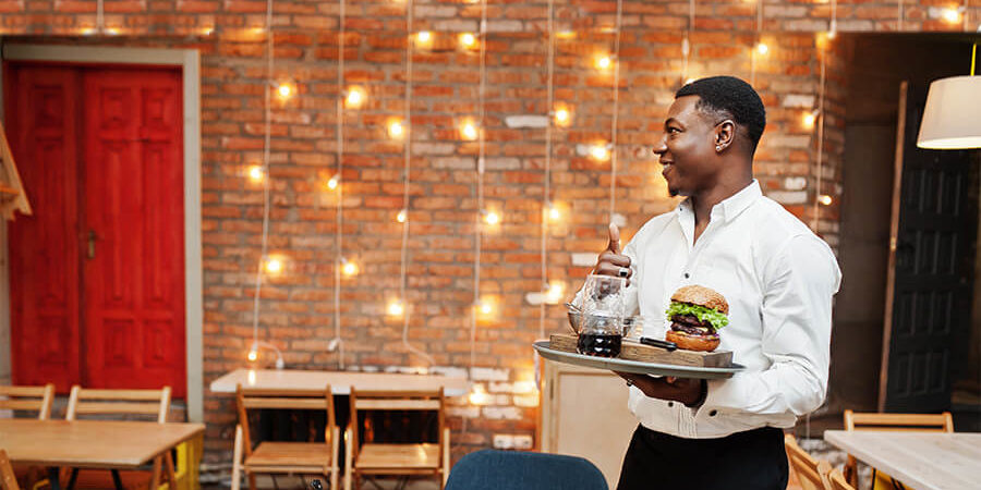 A waiter in a white shirt holding a tray with a burger and drink in a restaurant setting. - International Hotel School