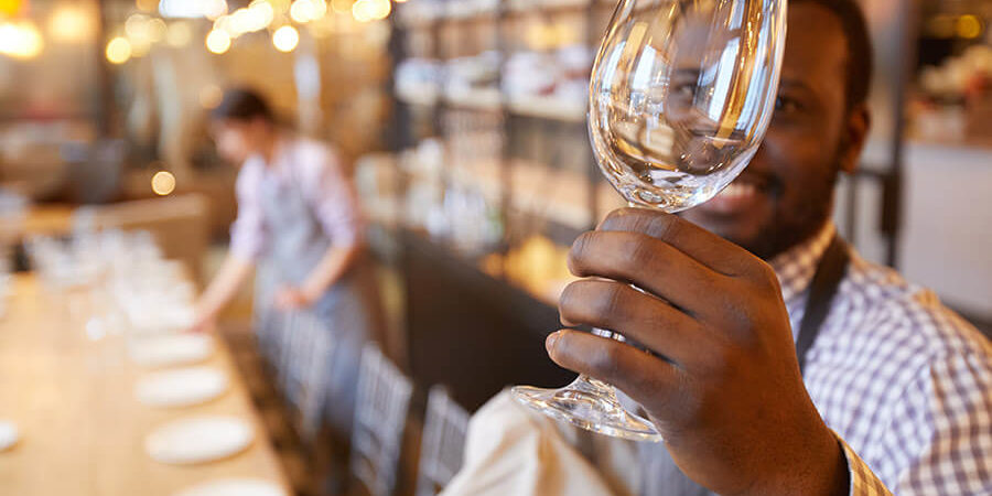 A waiter in an apron holding a clear wine glass in a restaurant setting. - International Hotel School