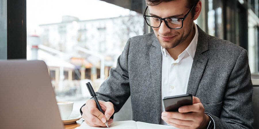 Man in a gray blazer writing in a notebook while using a smartphone and laptop at a table. - International Hotel School