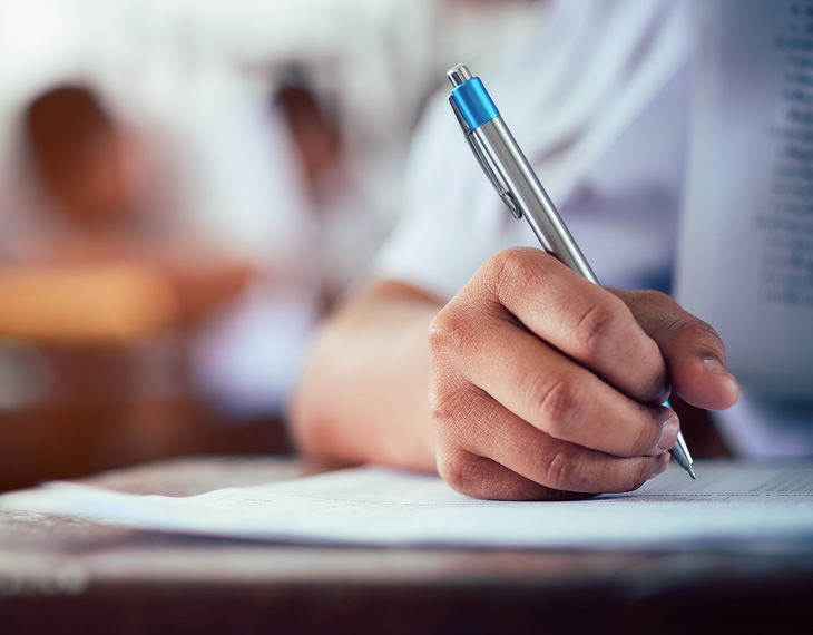 A student's hand holding a pen while writing on a piece of paper in a classroom setting. - International Hotel School