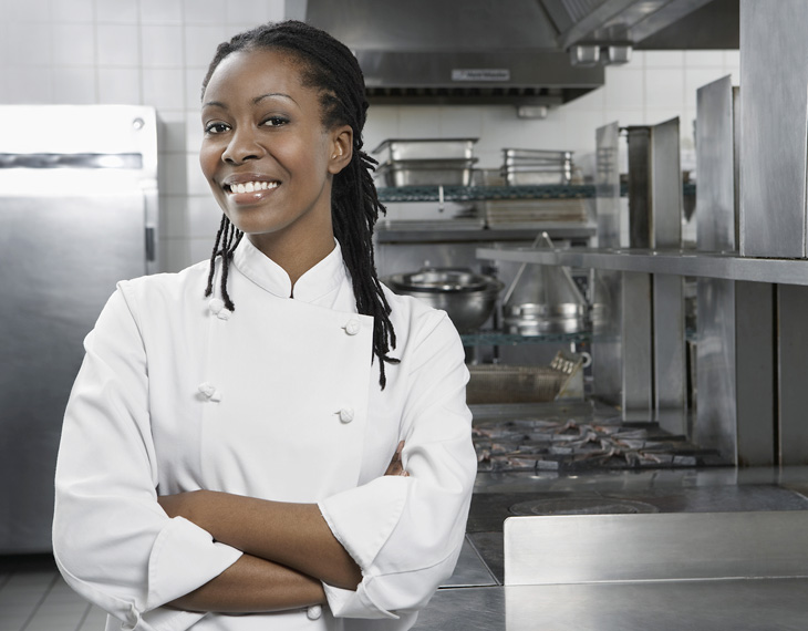Smiling female chef wearing a white uniform with arms crossed in a professional kitchen setting. - International Hotel School