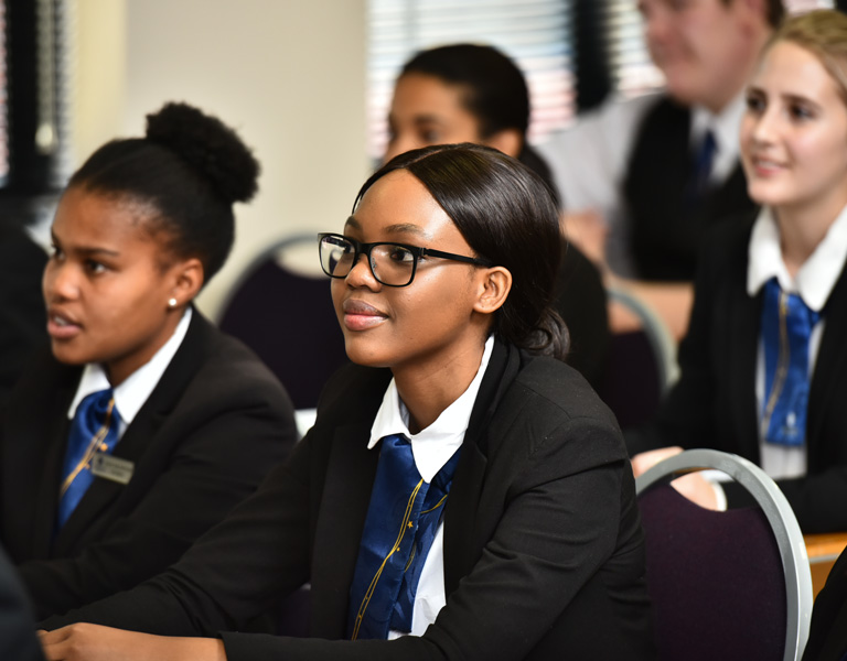 Group of students attentively listening in a classroom, with one student wearing glasses and a black blazer. - International Hotel School