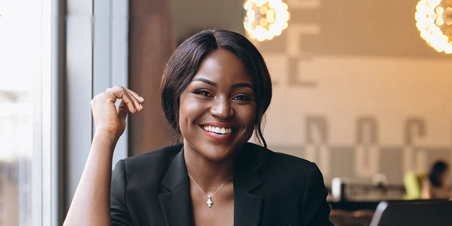 A woman in a black blazer smiling while sitting at a table with a laptop and a drink. - International Hotel School