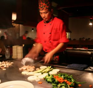 Chef in a red uniform skillfully preparing vegetables and shrimp at a teppanyaki grill. - International Hotel School