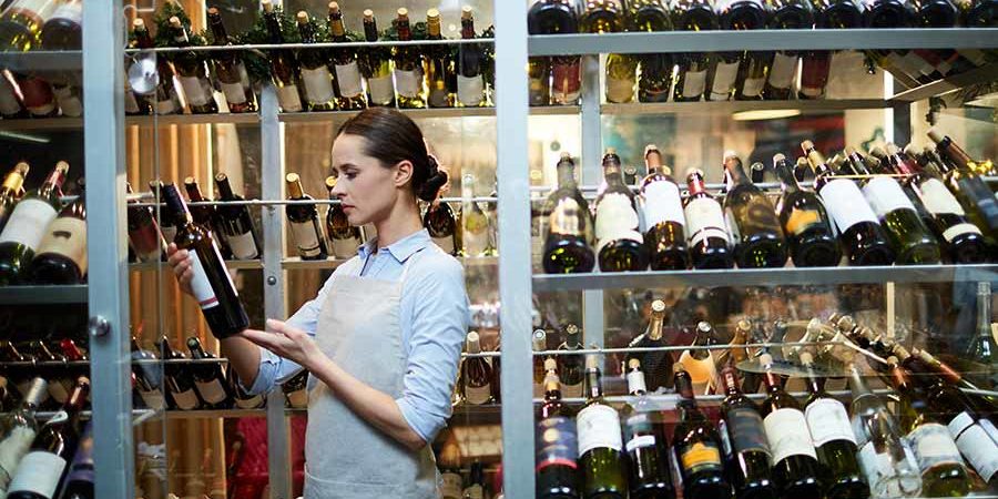 A woman in an apron examining a wine bottle in a glass display filled with various wine bottles. - International Hotel School