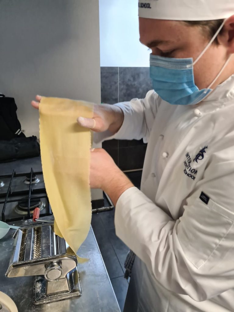 Chef in a white uniform and mask rolling out fresh pasta at a kitchen counter. - International Hotel School