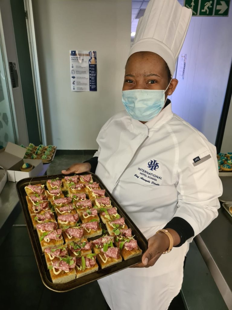 Chef in a white uniform presenting a tray filled with canapés at a culinary school. - International Hotel School