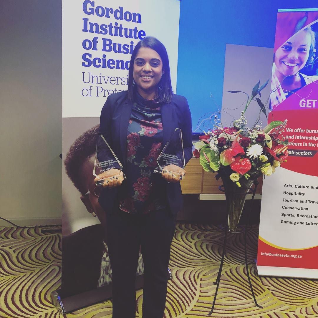 Individual holding two awards in front of a floral arrangement and banners at an event. - International Hotel School
