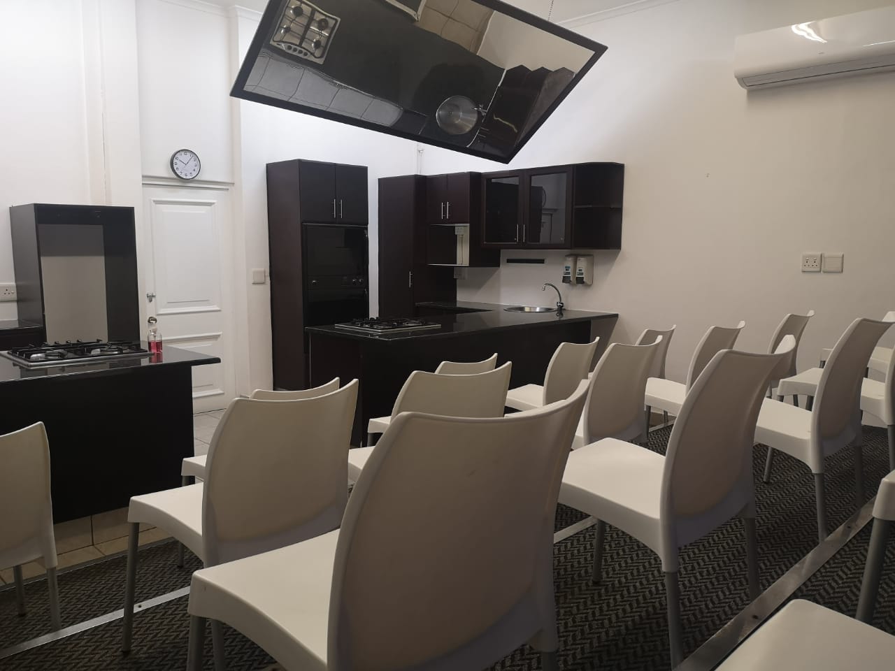 A modern kitchen with a cooking area and white chairs arranged for a demonstration or class. - International Hotel School