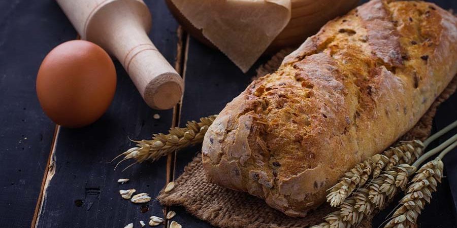 Loaf of freshly baked bread surrounded by baking ingredients including flour, an egg, and wheat. - International Hotel School