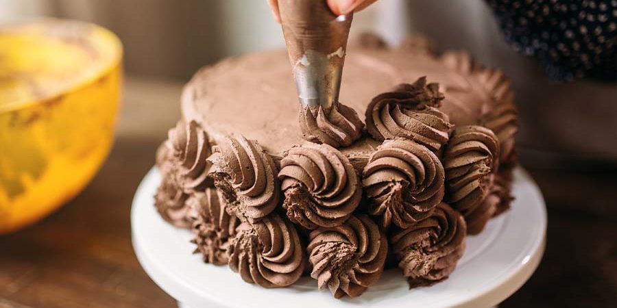Person decorating a chocolate cake with chocolate frosting using a piping bag. - International Hotel School