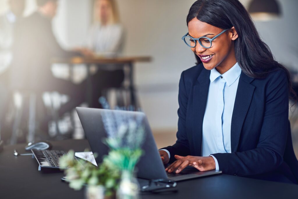 A professional woman in a suit using a laptop at a desk with plants in the foreground. - International Hotel School