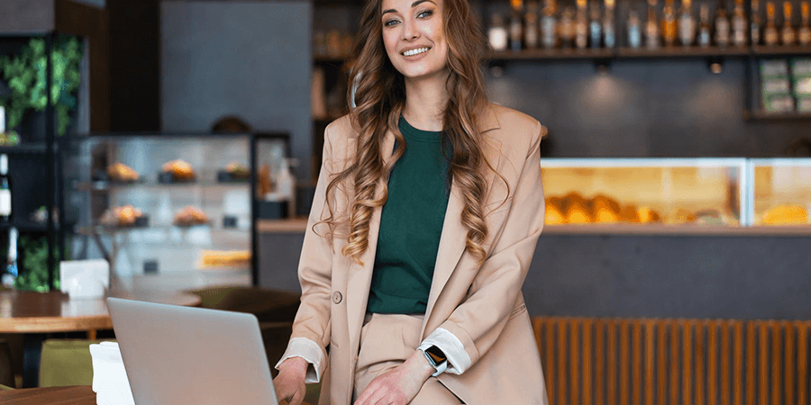 A woman in a beige suit sitting at a table with a laptop in a modern cafe setting. - International Hotel School