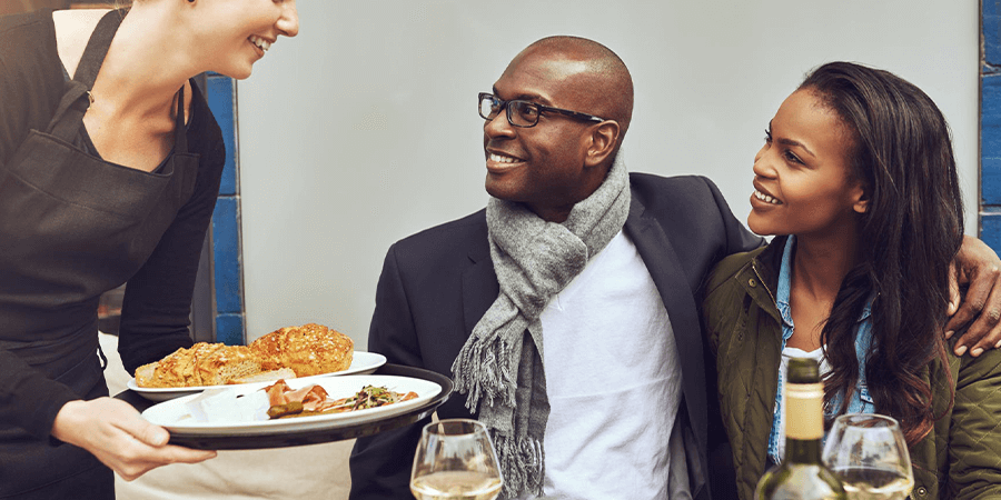 A waitress serving a plate of food to a couple seated at a table with wine glasses. - International Hotel School