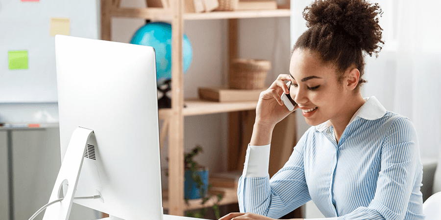 A woman in a blue striped shirt sitting at a desk using a computer and talking on the phone. - International Hotel School
