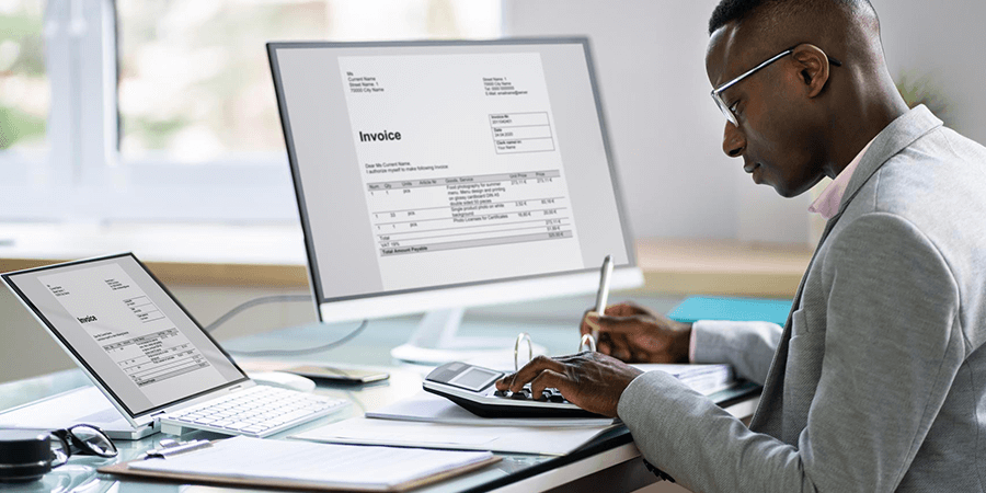 A man in a suit reviewing invoices on a computer and tablet at a modern desk. - International Hotel School