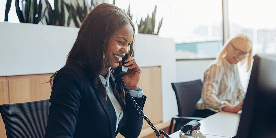 A woman in a business suit talking on the phone at a desk in an office environment. - International Hotel School
