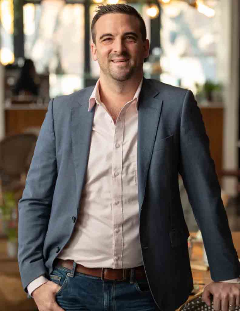 A man wearing a light pink shirt and a dark blazer, standing confidently with a smile. - International Hotel School