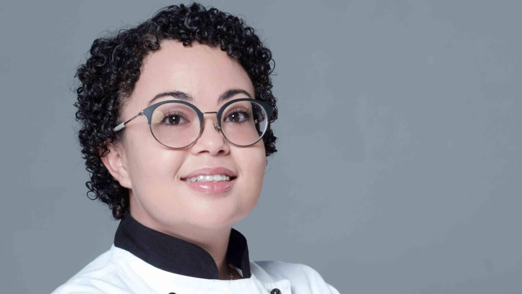 Chef wearing a white uniform and glasses, smiling against a gray background. - International Hotel School