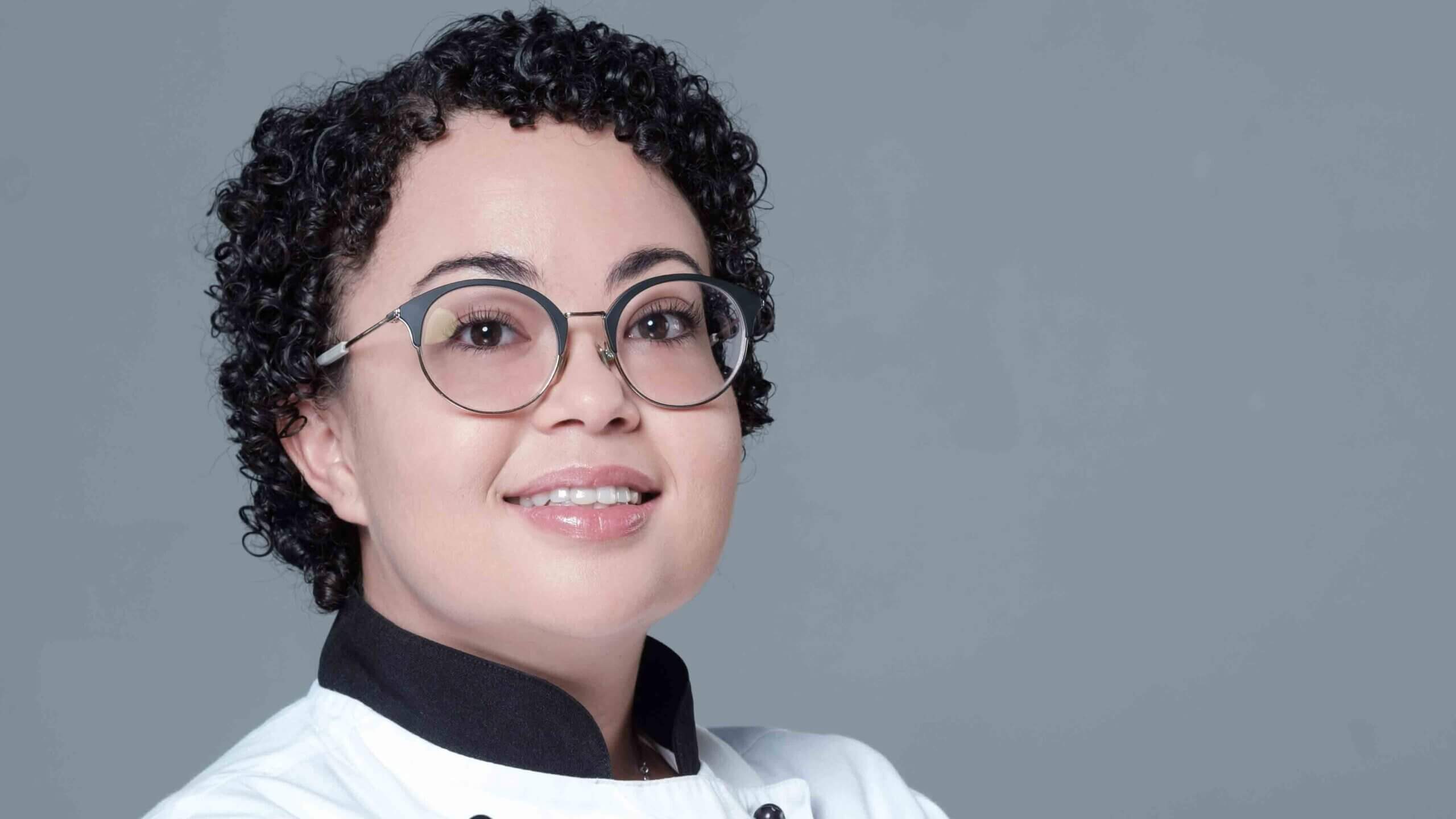 Chef wearing a white uniform and glasses, smiling against a gray background. - International Hotel School