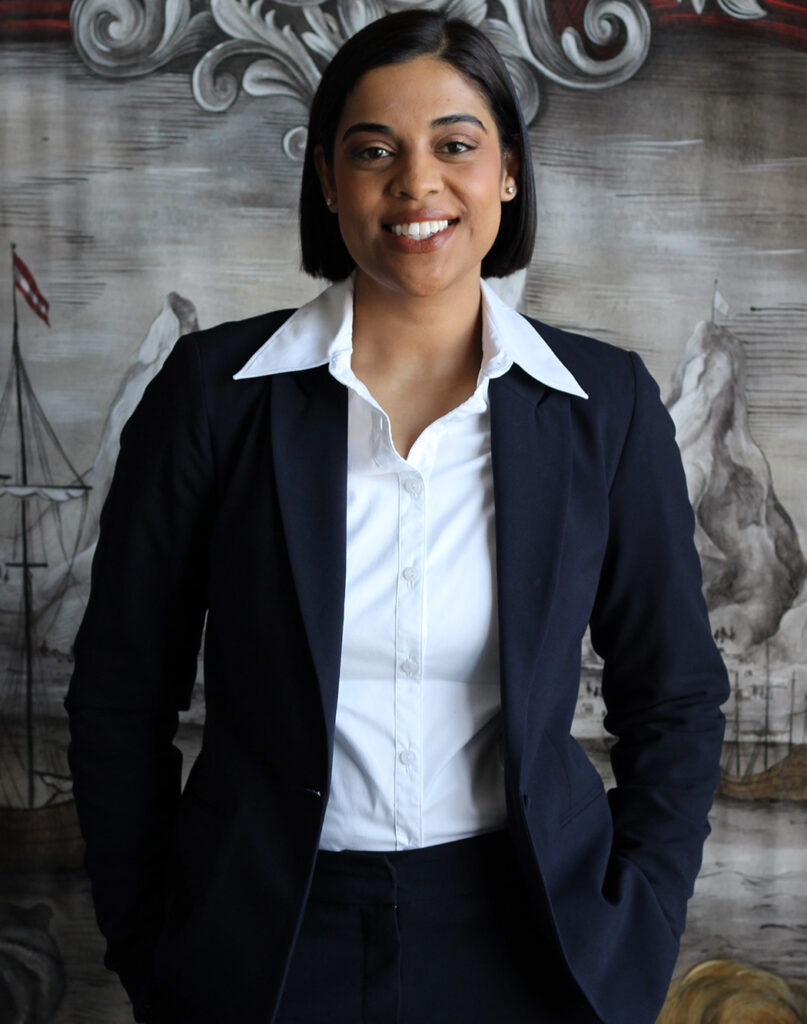 A woman in a black suit and white shirt smiling confidently in front of a decorative background. - International Hotel School