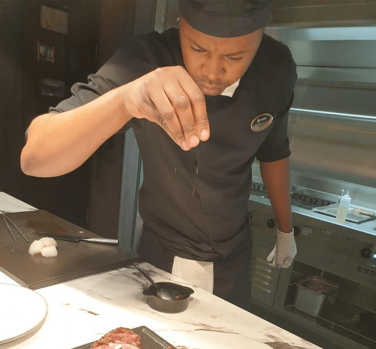 Chef in a black uniform seasoning a dish in a professional kitchen environment. - International Hotel School
