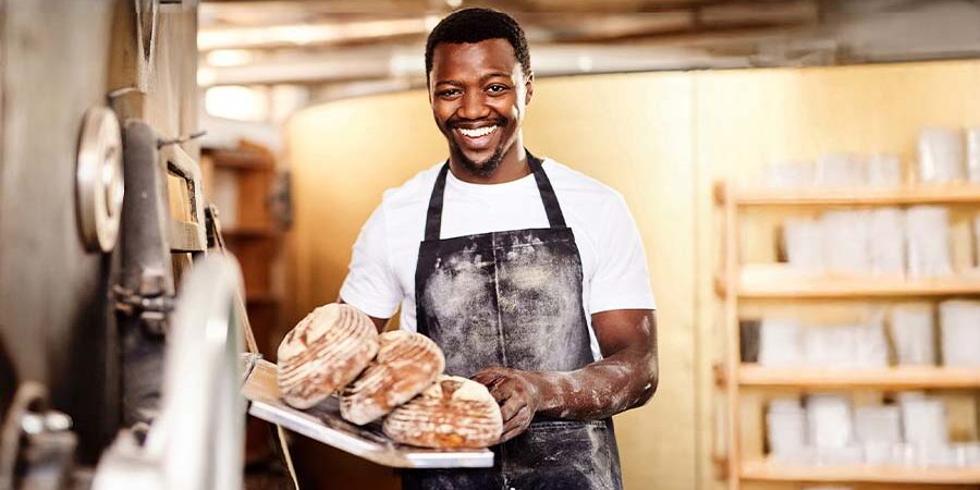 A smiling assistant baker wearing an apron, holding a tray of freshly baked bread in a bakery. - International Hotel School