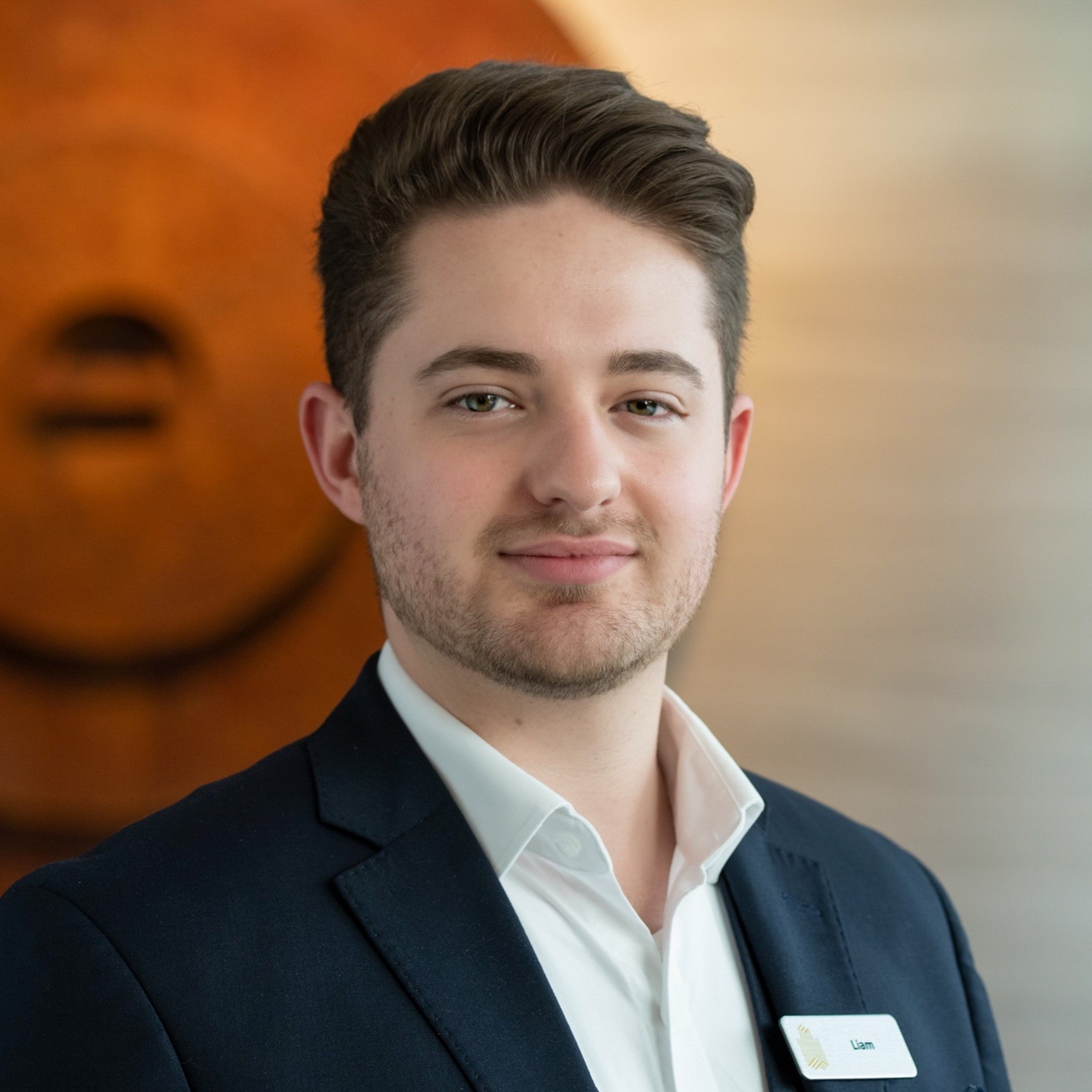 Young man in a dark suit and white shirt with a name tag, posing for a professional headshot. - International Hotel School
