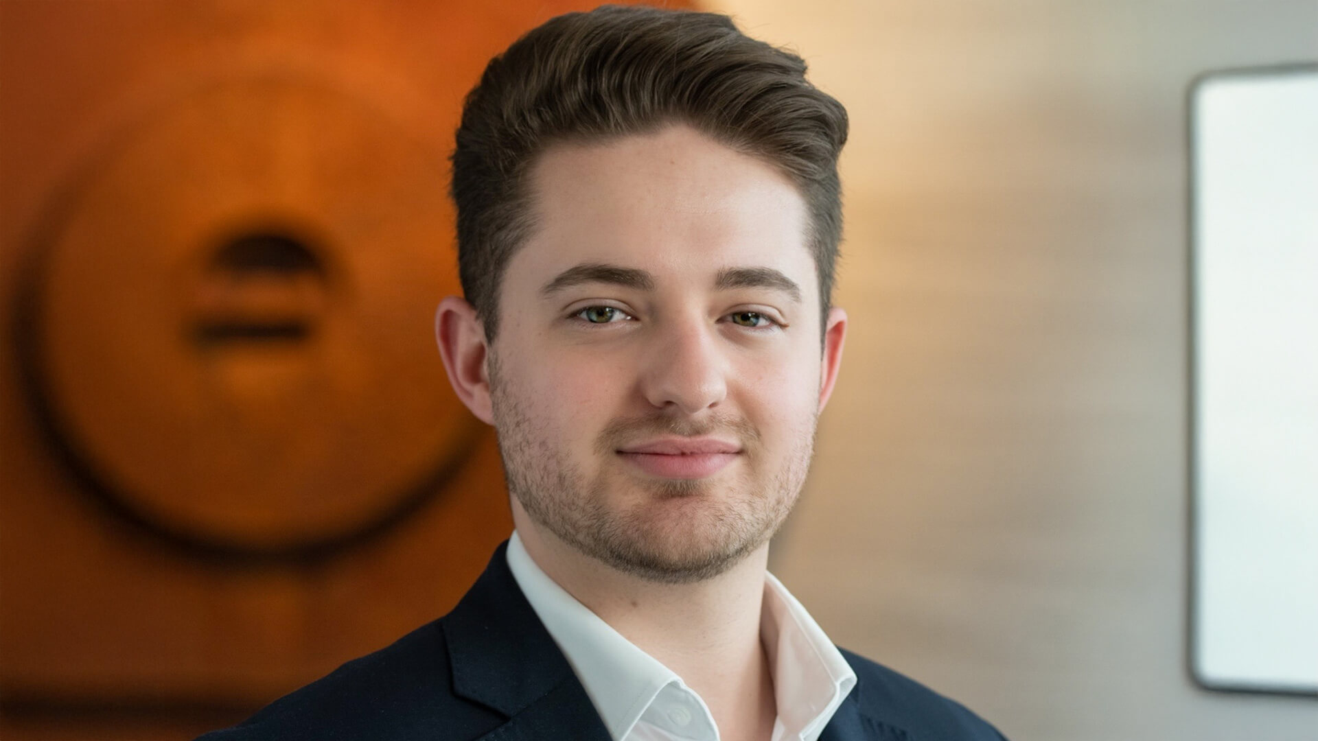 Young man in a dark suit with a white shirt, smiling against a neutral background. - International Hotel School