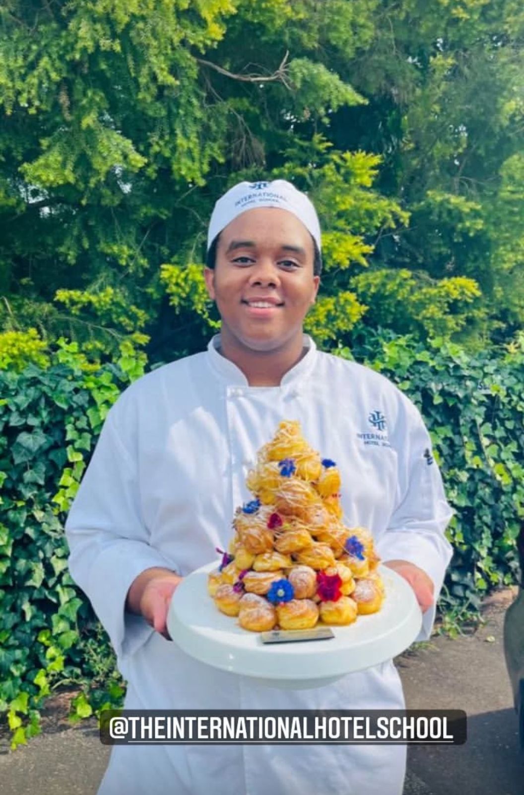 A pastry chef in a white uniform presenting a platter of desserts decorated with flowers. - International Hotel School