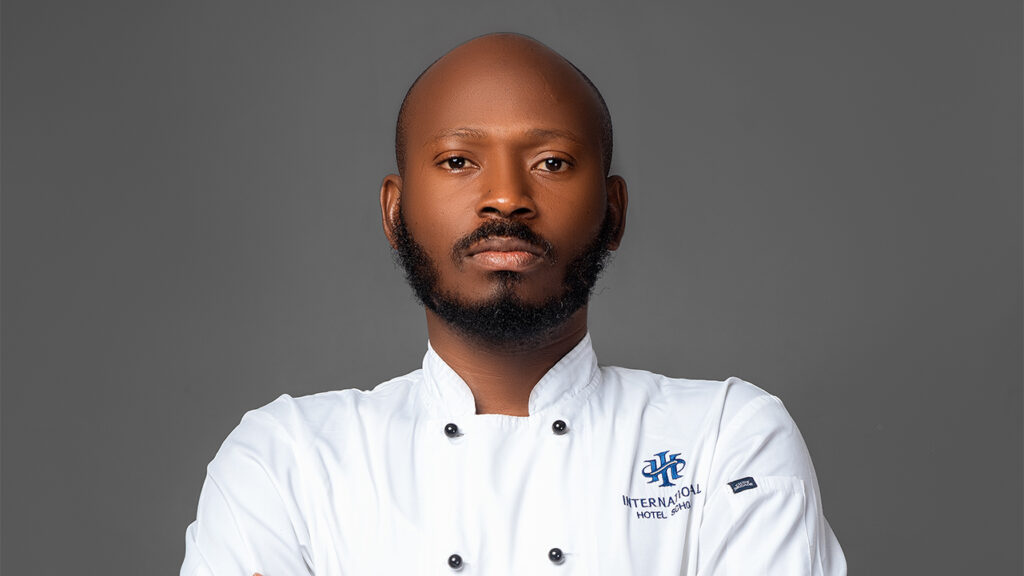 A chef wearing a white culinary uniform with a logo, arms crossed and a serious expression. - International Hotel School